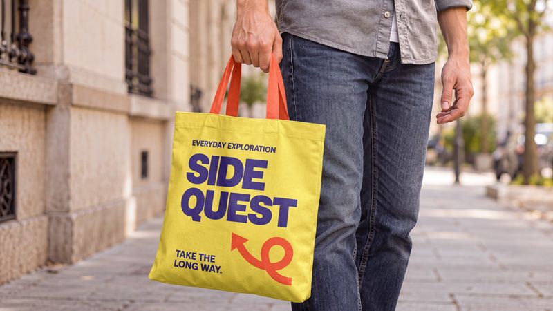 Canvas tote bag mockup in hand with yellow fabric, orange handles, and purple lettering on a city sidewalk