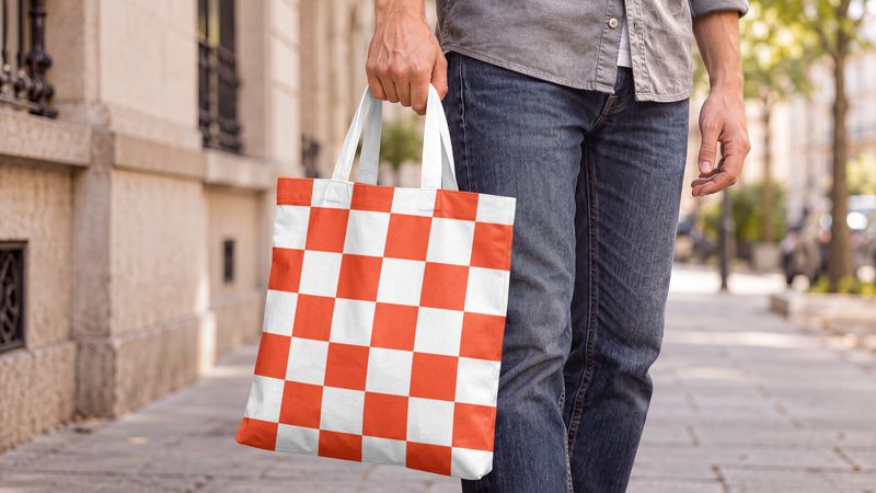 Canvas tote bag mockup in hand with an orange and white checkerboard print and white handles